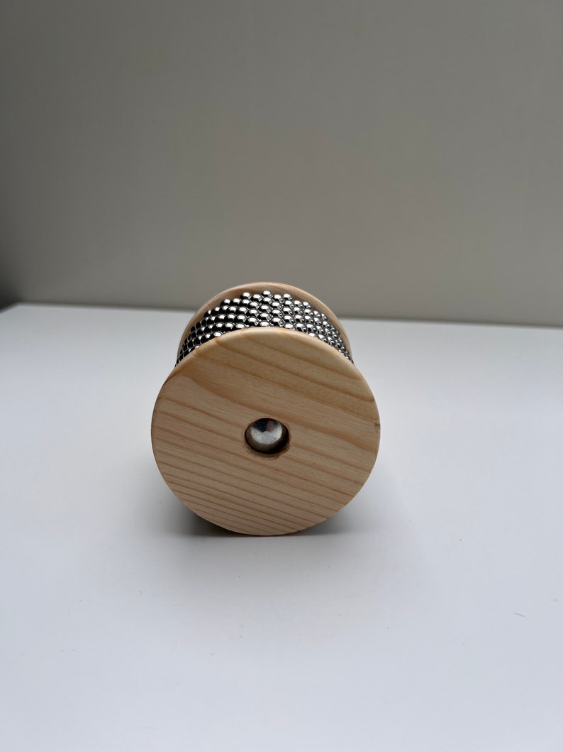 May include: A wooden percussion instrument with a circular shape and a band of metal beads around the center. The instrument is made of light wood and has a natural finish.