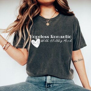 Może przedstawiać: Szary t-shirt z nadrukiem "Hopeless Romantic With A Filthy Mind" i białym sercem. Modelka nosi niebieskie dżinsy i białe buty sportowe.