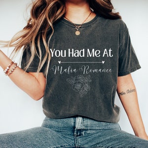 Op de afbeelding: Een donkergrijs T-shirt met de tekst "You Had Me At Mafia Romance" in witte letters. De tekst wordt omringd door twee kleine hartjes en een bloemmotief.
