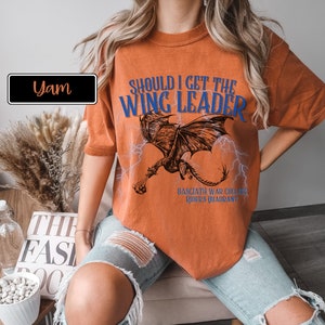 K&ouml;nnte beinhalten: Orangefarbenes T-Shirt mit einem Drachen-Motiv und dem Text "Should I get the Wing Leader" in Blau. Der Text "Basciath War College, Rider's Quadrant" ist unter dem Drachen gedruckt.