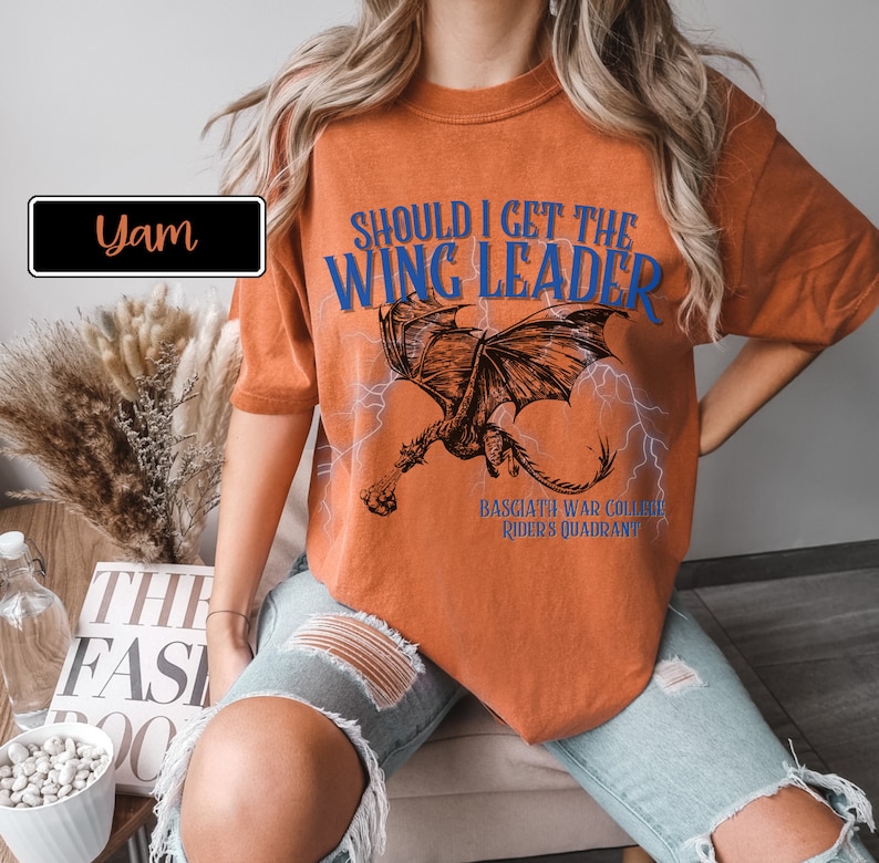 K&ouml;nnte beinhalten: Orangefarbenes T-Shirt mit einem Drachen-Motiv und dem Text "Should I get the Wing Leader" in Blau. Der Text "Basciath War College, Rider's Quadrant" ist unter dem Drachen gedruckt.
