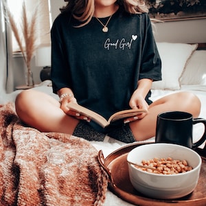 以下が含まれることがあります： 「グッドガール」という文字と小さなハートが書かれた黒いTシャツを着た女性。彼女はベッドに座って、膝の上に本を置き、目の前にトレイにマグカップとシリアルボウルを置いている。