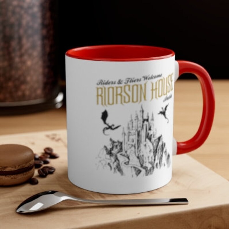 May include: White ceramic mug with a red rim and handle. The mug features a black and white illustration of a castle with dragons and the text "Riders & Fliers Welcome Riorson House".