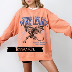 May include: Terracotta orange sweatshirt with a graphic of a dragon flying with lightning bolts and the text "Should I get the Wing Leader" and "Basgiath War College Rider's Quadrant".