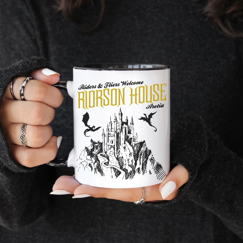 May include: White ceramic mug with a black rim. The mug features a black and white illustration of a castle with two dragons flying above it. The text "Riders & Fliers Welcome Riorson House Aretia" is printed in gold on the mug.