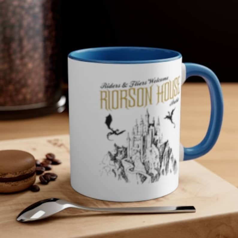 May include: A white ceramic mug with a blue rim and handle. The mug features a black and white illustration of a castle with dragons flying overhead. The text "Riders & Fliers Welcome RIORSON HOUSE" is printed in gold on the mug.