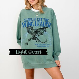 May include: A light green sweatshirt with the text "Should I get the Wing Leader" in blue and black. A black and white illustration of a dragon with lightning bolts is printed on the front. The text "Basgiath War College Rider's Quadrant" is printed below the dragon.