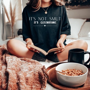 May include: A woman wearing a black t-shirt with the text "It's not smut it's cliterature" in white lettering. She is sitting on a bed with a book in her lap and a mug and bowl of cereal on a tray in front of her.