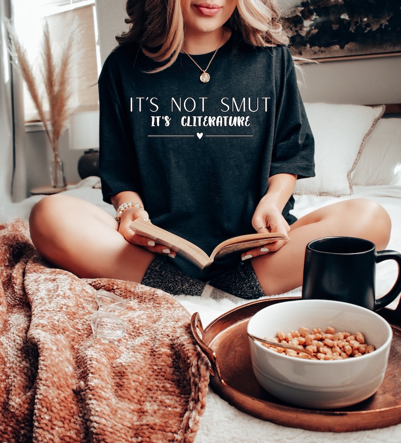 May include: A woman wearing a black t-shirt with the text "It's not smut it's cliterature" in white lettering. She is sitting on a bed with a book in her lap and a mug and bowl of cereal on a tray in front of her.
