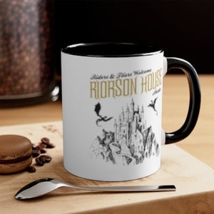 May include: White ceramic mug with a black rim and handle. The mug has a black and white illustration of a castle with dragons and the text "Riders & Fliers Welcome RIORSEN HOUSE".