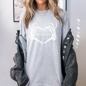 May include: Gray t-shirt with a white skeleton hand heart design and the text "skeletally" printed on the front.