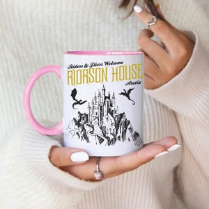 May include: A white ceramic mug with a pink rim and handle. The mug features a black and white illustration of a castle with two dragons flying above it. The text "Riders & Fliers Welcome Riorson House Aretia" is printed in gold on the mug.