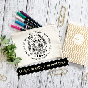 Può includere: Una pochette con cerniera in tela bianca con un design in bianco e nero che presenta due scheletri che si tengono per mano con il testo "I still read fairytales, they are just darker now". La pochette è su una superficie di legno bianco con graffette dorate e un piccolo taccuino con un motivo dorato e bianco.
