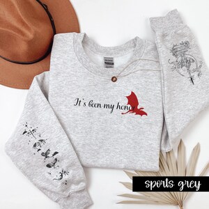 May include: A grey sweatshirt with the text "It's been my honor" and a red dragon graphic. The sleeves have black dragon graphics. The sweatshirt is available in sports grey.