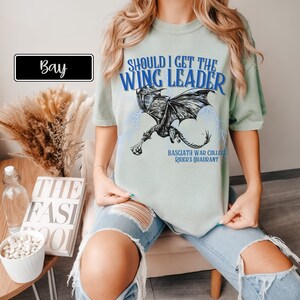 K&ouml;nnte beinhalten: Ein hellblaues T-Shirt mit einem schwarzen Drachen-Motiv und dem Text "Should I get the Wing Leader" in Blau. Der Text "Basgiath War College Rider's Quadrant" ist ebenfalls in Blau auf dem T-Shirt aufgedruckt.