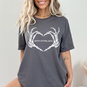 May include: Gray t-shirt with a white skeleton hand design making a heart shape. The text "STFUATTDLAGG" is inside the heart.
