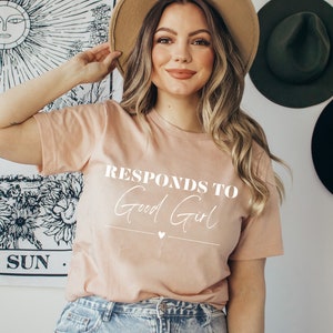 May include: A light pink t-shirt with the text "Responds to Good Girl" in white lettering. The shirt has a small heart symbol below the text.
