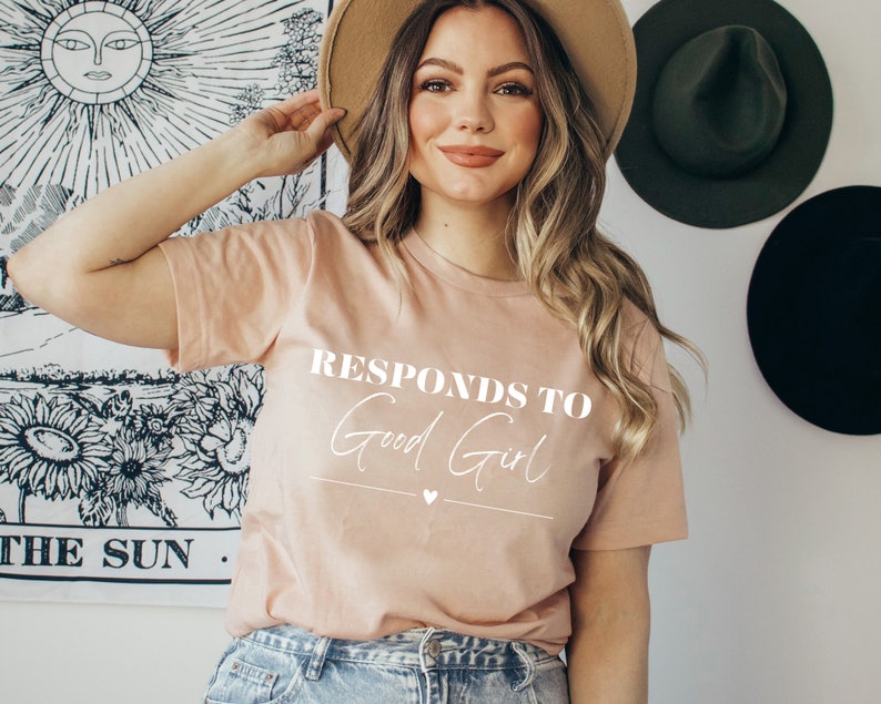 May include: A light pink t-shirt with the text "Responds to Good Girl" in white lettering. The shirt has a small heart symbol below the text.