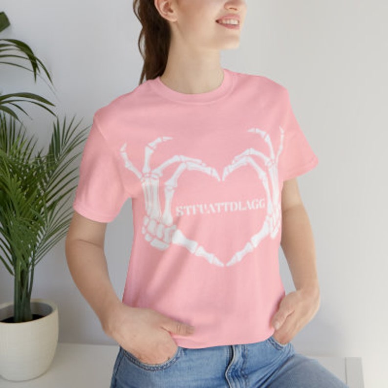 May include: Pink t-shirt with a white skeleton hand design making a heart shape. The text "STRATTOLAGG" is inside the heart.