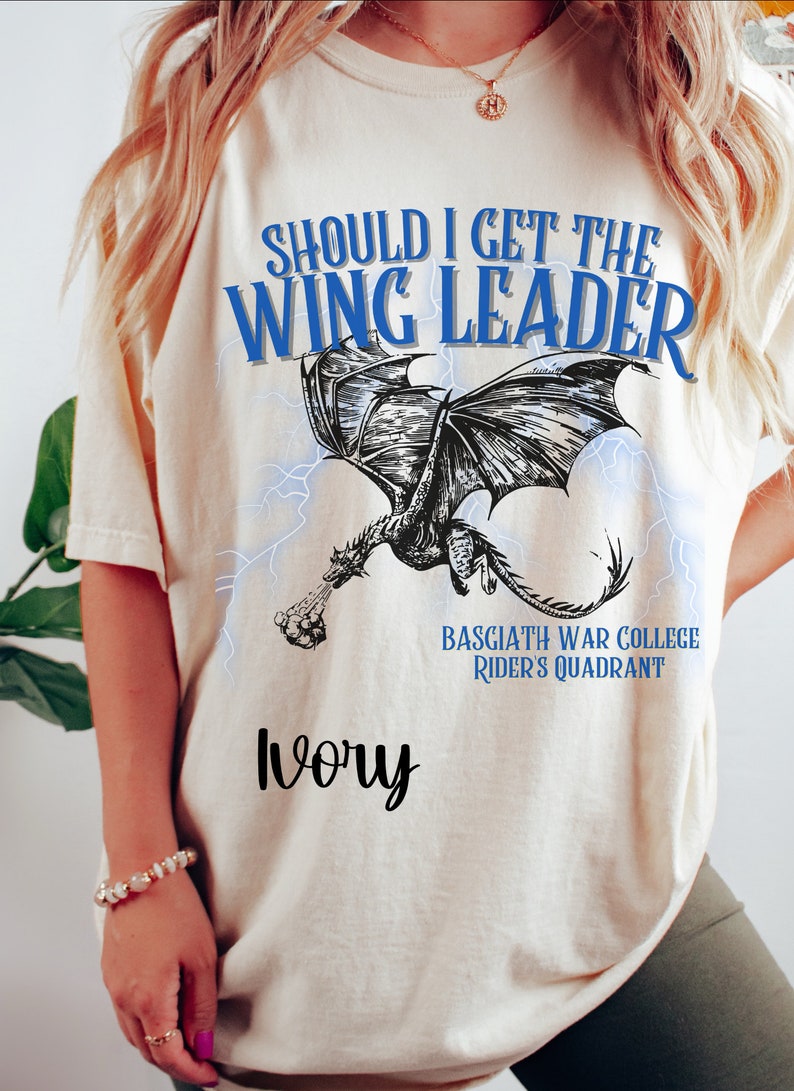 K&ouml;nnte beinhalten: Ein cremefarbenes T-Shirt mit einer blauen Grafik eines Drachen mit Blitzen. Der Text "Should I get the Wing Leader" befindet sich &uuml;ber dem Drachen und "Basgiath War College Rider's Quadrant" unter dem Drachen. Das Wort "Ivory" ist in schwarzer Kursivschrift unter der Grafik geschrieben.