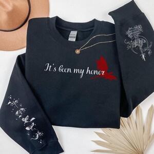 May include: Black sweatshirt with the text "It's been my honor" and a red bird graphic on the front. The sleeves have white graphic designs of birds and stars.