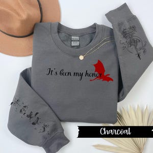 May include: A charcoal grey sweatshirt with a red dragon graphic and the text "It's been my honor". The sleeves have a black and white graphic of a dragon with a sword and a moon.