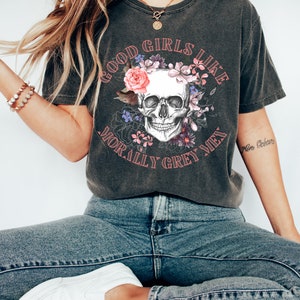 May include: A dark grey t-shirt featuring a skull graphic surrounded by flowers. The text "Good Girls Like Morally Grey Men" is printed on the shirt.