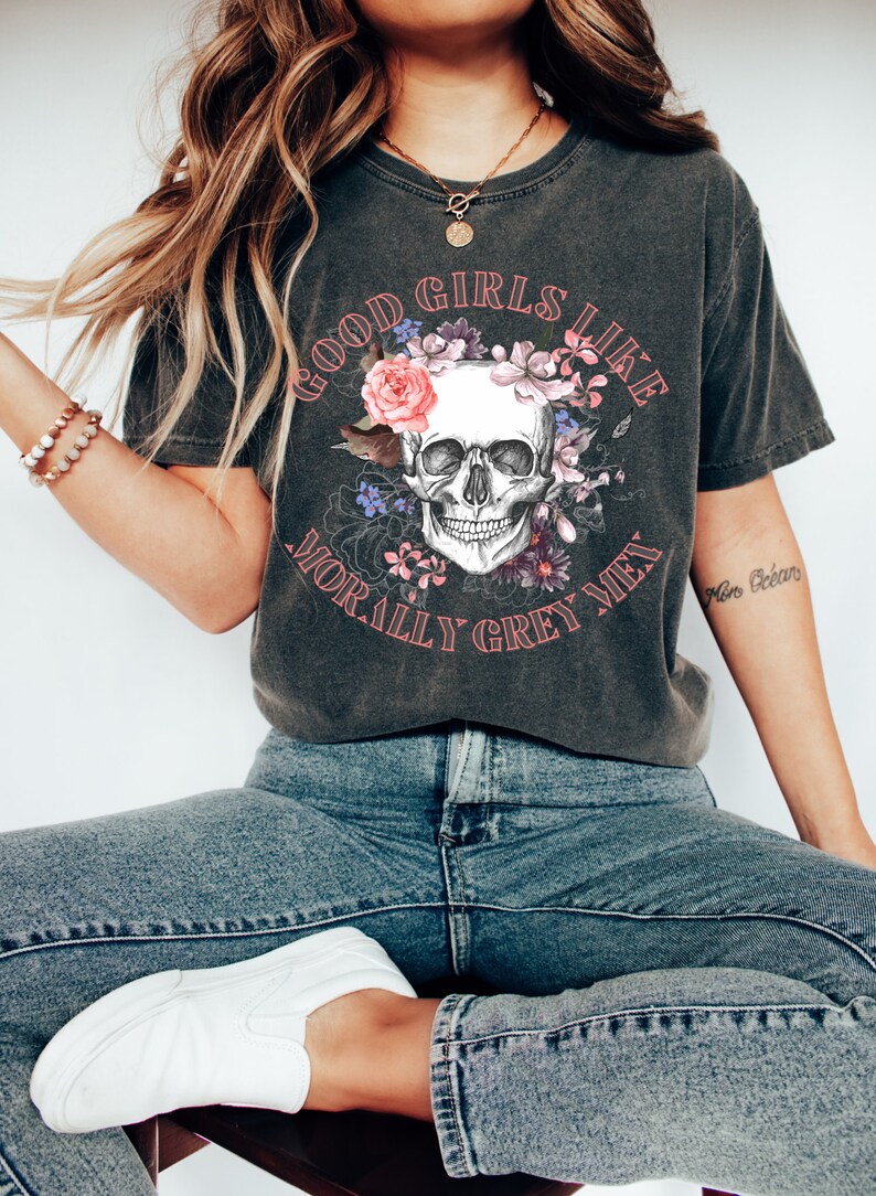 May include: A dark grey t-shirt featuring a skull graphic surrounded by flowers. The text "Good Girls Like Morally Grey Men" is printed on the shirt.