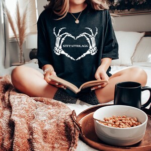May include: A black t-shirt with a white skeleton hand design making a heart shape. The text "STFUATTLDAGG" is printed inside the heart.