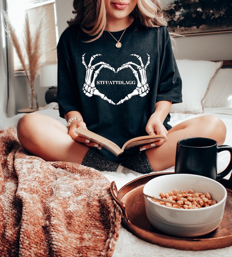 May include: A black t-shirt with a white skeleton hand design making a heart shape. The text "STFUATTLDAGG" is printed inside the heart.