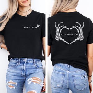 K&ouml;nnte beinhalten: Schwarzes T-Shirt mit dem Text "GOOD GIRL" und einem Herz auf der Vorderseite. Die R&uuml;ckseite des Shirts zeigt eine Skelett-Hand-Herzform mit dem Text "SITUATTDLAGG".