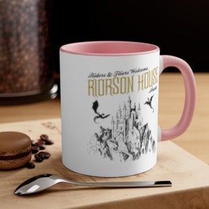 May include: A white ceramic mug with a pink rim and handle. The mug features a black and white illustration of a castle with dragons flying overhead. The text "Riders & Fliers Welcome RIORSON HOUSE" is printed in gold on the mug.