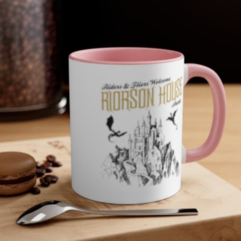 May include: A white ceramic mug with a pink rim and handle. The mug features a black and white illustration of a castle with dragons flying overhead. The text "Riders & Fliers Welcome RIORSON HOUSE" is printed in gold on the mug.