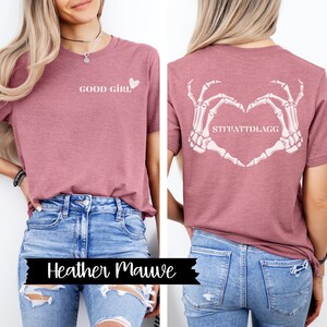 K&ouml;nnte beinhalten: Ein T-Shirt in Heide-Mauve mit dem Text "GOOD GIRL" und einem kleinen Herz auf der Vorderseite. Die R&uuml;ckseite des Shirts zeigt ein Skelett-Hand-Herz-Design mit dem Text "SITUATTDLAGG".