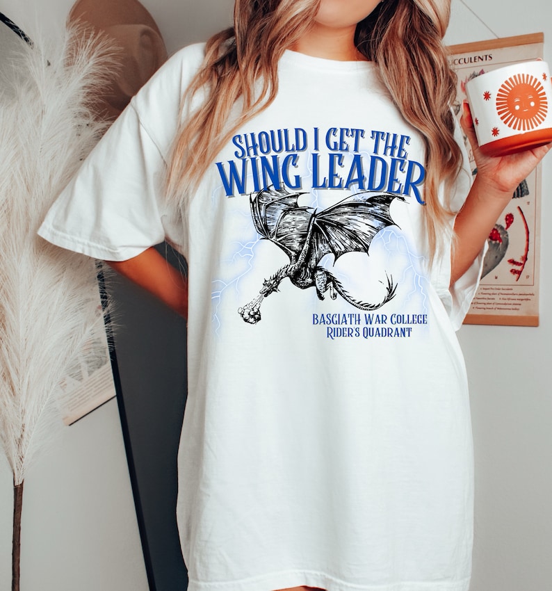K&ouml;nnte beinhalten: Wei&szlig;es T-Shirt mit einem schwarzen und wei&szlig;en Drachen-Motiv und blauem Text, der "Should I get the Wing Leader" und "Basciath War College Rider's Quadrant" lautet.