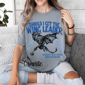 K&ouml;nnte beinhalten: Graues T-Shirt mit einer blauen Grafik eines Drachen mit Blitzen und dem Text "Should I get the Wing Leader" und "Basciath War College Riders Quadrant".