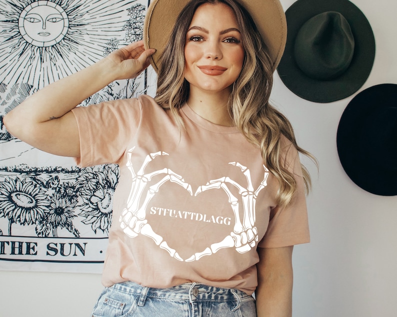 May include: A light pink t-shirt with a white skeleton hand design making a heart shape. The text "STFUATTDLAGG" is inside the heart.