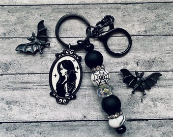 May include: A black keychain featuring a black and white beaded charm, an oval pendant with a woman and raven design, and two black bat charms. The keychain has a black clasp and key ring.