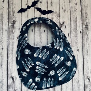 May include: A dark blue baby bib with a white Halloween-themed pattern of witches, potion bottles, and brooms. The bib has a rounded shape with a neck opening and a snap closure. Two black bat cutouts are in the background.
