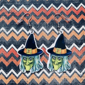 May include: A pair of Halloween-themed earrings featuring a witch's face and hat. The witch has green skin, a crooked nose, and blue hair. The hat is black with an orange band and buckle. The earrings hang from silver-colored hooks.