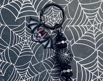 May include: A black and white bat charm with red eyes hangs from a black metal key ring. The charm is attached to a black beaded chain with a black and white spiderweb bead.
