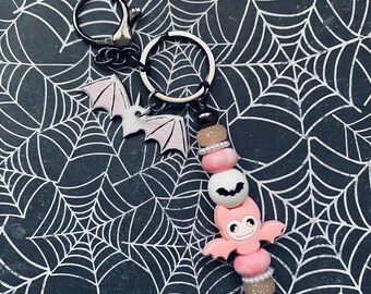 May include: A pink and white bat-themed keyring with a silver key ring. The keyring features a pink bat charm with a white face and black eyes, and a white bat charm with pink accents. The keyring is strung with pink and white beads.