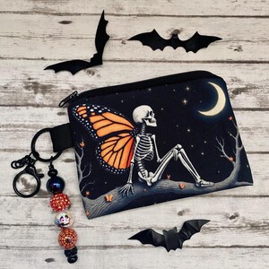 Peut inclure: Une pochette zippée noire illustrée d'un squelette aux ailes de papillon monarque orange, assis sur une branche sous un croissant de lune et des étoiles. Un porte-clés noir est attaché, orné de perles dont un crâne, une toile d'araignée et des paillettes.