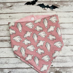 Może przedstawiać: Różowa bandana z białym wzorem duchów. Bandana ma zapięcie na zatrzask i jest dwustronna z miękką różową stroną z polaru.