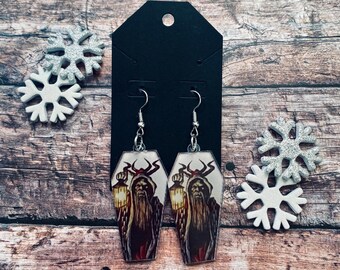 May include: A pair of coffin-shaped earrings featuring a spooky illustration of a figure holding a lantern. The earrings are silver-toned and hang from a black card. Decorative snowflakes are on either side.