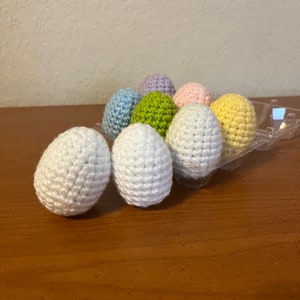 May include: A set of eight crocheted Easter eggs in a variety of pastel colors, including white, light blue, light green, light pink, and yellow. The eggs are arranged in a plastic egg carton.