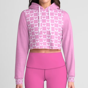 May include: A pink cropped hoodie with a white checkered pattern featuring hearts. The hoodie has long sleeves and a drawstring hood.