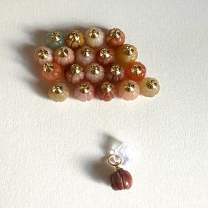 May include: A collection of small, pumpkin-shaped beads in various colors, including orange, pink, and yellow, each with a gold-colored cap. One bead is attached to a gold-colored ring. The beads are arranged on a white surface.