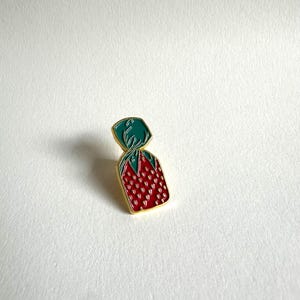 May include: A gold-colored enamel pin shaped like a wrapped candy with a red and white polka dot pattern and a green wrapper.