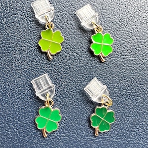 May include: Four pairs of clover earrings. Each earring features a bright green, four-leaf clover charm with gold-colored accents. The earrings have clear, plastic ear posts.